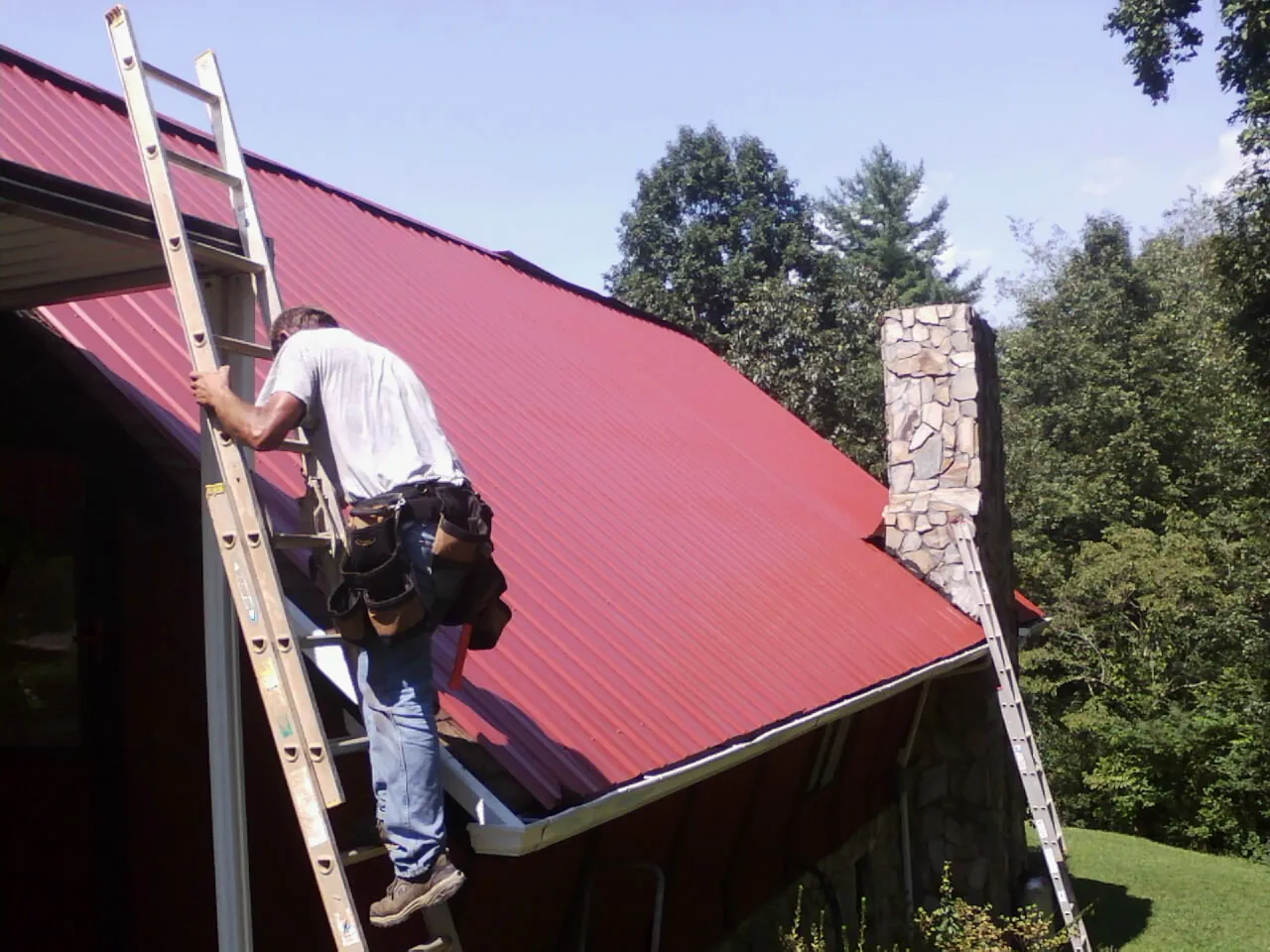 Roofer on a ladder beside a red metal roof and stone chimney — Soffit & Fascia Repair by Roofing Brothers in Shady Valley
